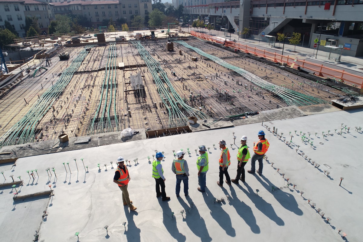 Looking down on a finished concrete deck: seven construction workers in high-visibility vests standing in a loose line, each casting a long shadow across the slab, with a rebar and conduit deck behind them.