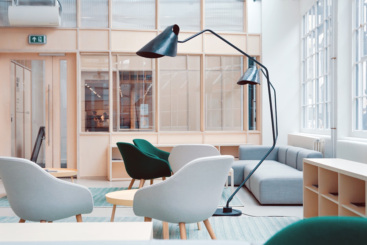 An empty lounge with low upholstered chairs, round side tables, a tall arc floor lamp and glazed partitions along the back wall. Nobody is in the room.