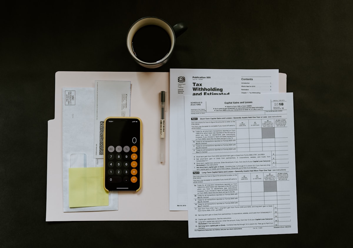 Overhead view of a desk: printed tax and variance paperwork spread out, a calculator, a pen resting on a page, and a mug of coffee.