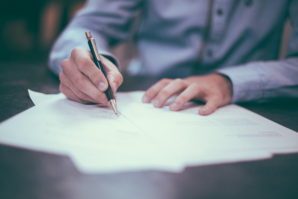Two hands over loose sheets of paper on a dark desk, one steadying the page and one holding a pen just touching the line, in soft diffuse light.