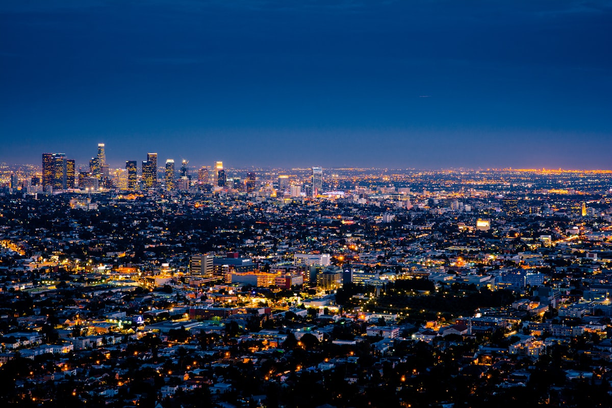 A city seen from above after dark: a dense field of lit windows and streetlights running back to a small downtown cluster on the horizon.