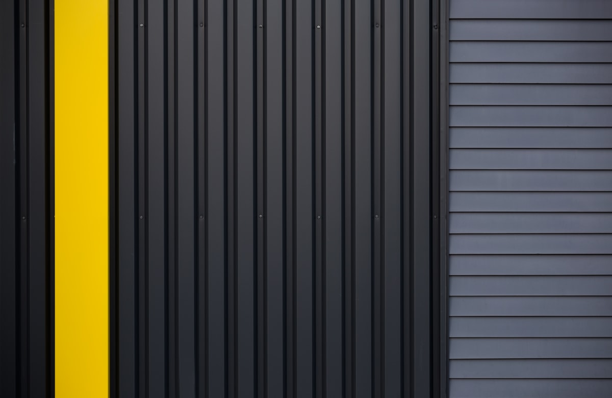 Dark ribbed metal cladding photographed flat on, one bay painted a single vertical stripe of yellow.